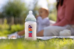 Babyflasche mit bunter Aufschrift 'HiPP' steht auf Gras im Vordergrund, unscharfe Eltern und Kind im Hintergrund