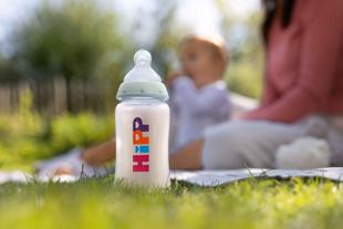 Babyflasche mit bunter Aufschrift 'HiPP' steht auf Gras im Vordergrund, unscharfe Eltern und Kind im Hintergrund