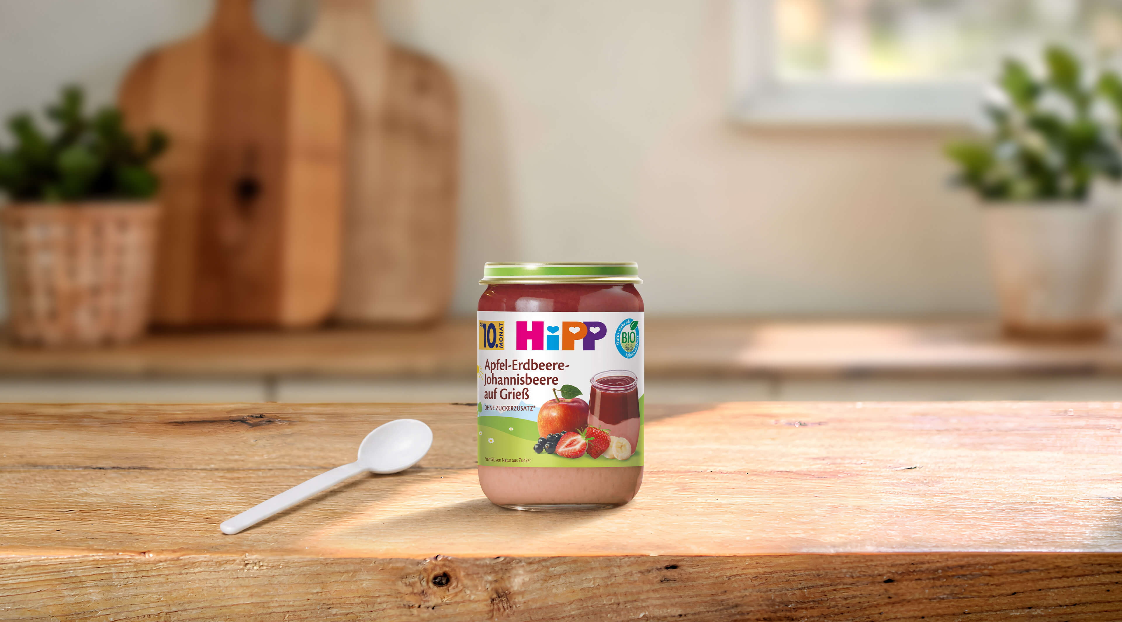 HiPP Bio-Gläschen Apfel-Erdbeere-Johannisbeere auf Grieß ab 10 Monaten auf Holztisch mit Löffel in heller Küche