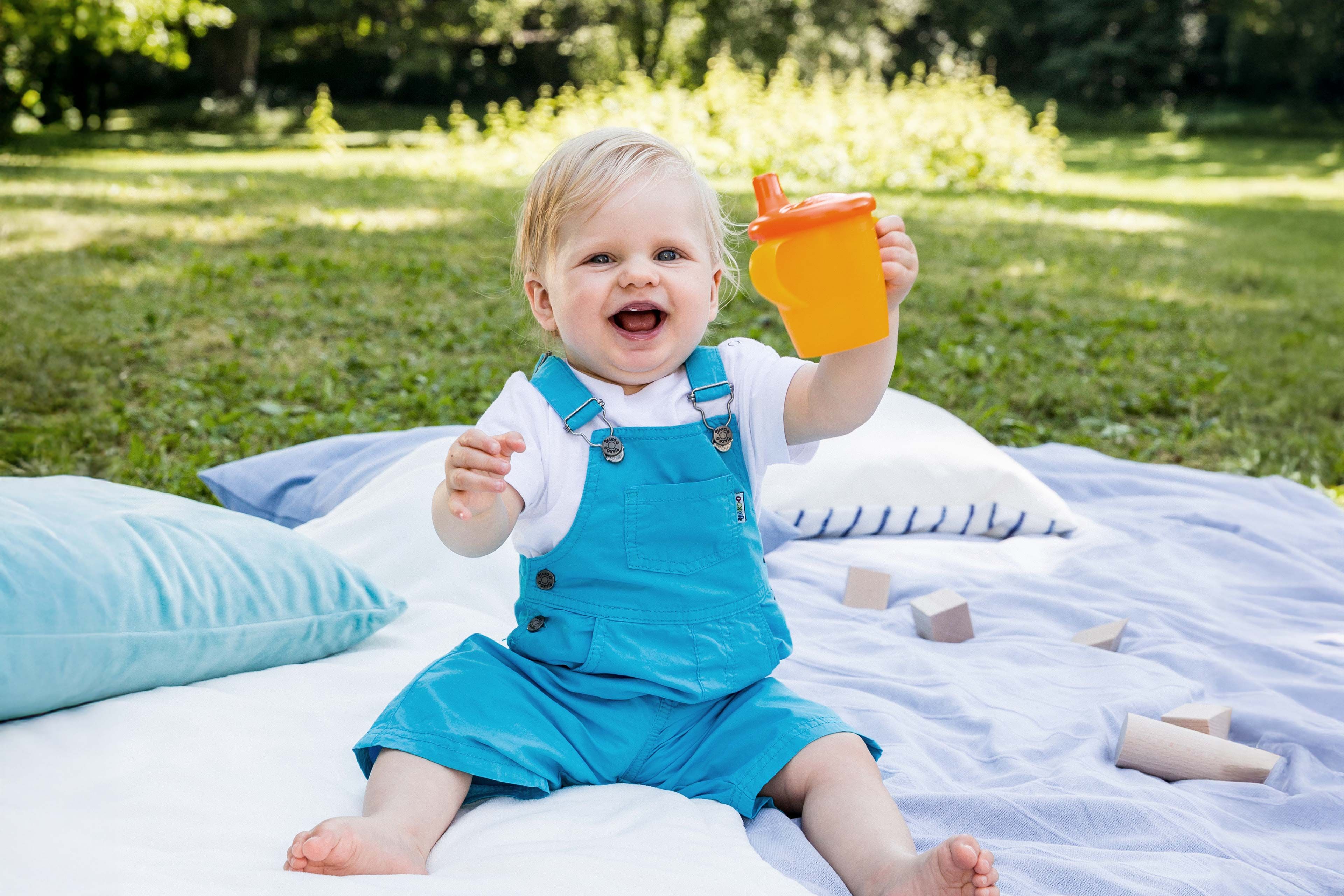 Lachendes Baby im Garten sitzt auf einer Decke und hält fröhlich einen Trinkbecher mit Babygetränk hoch.