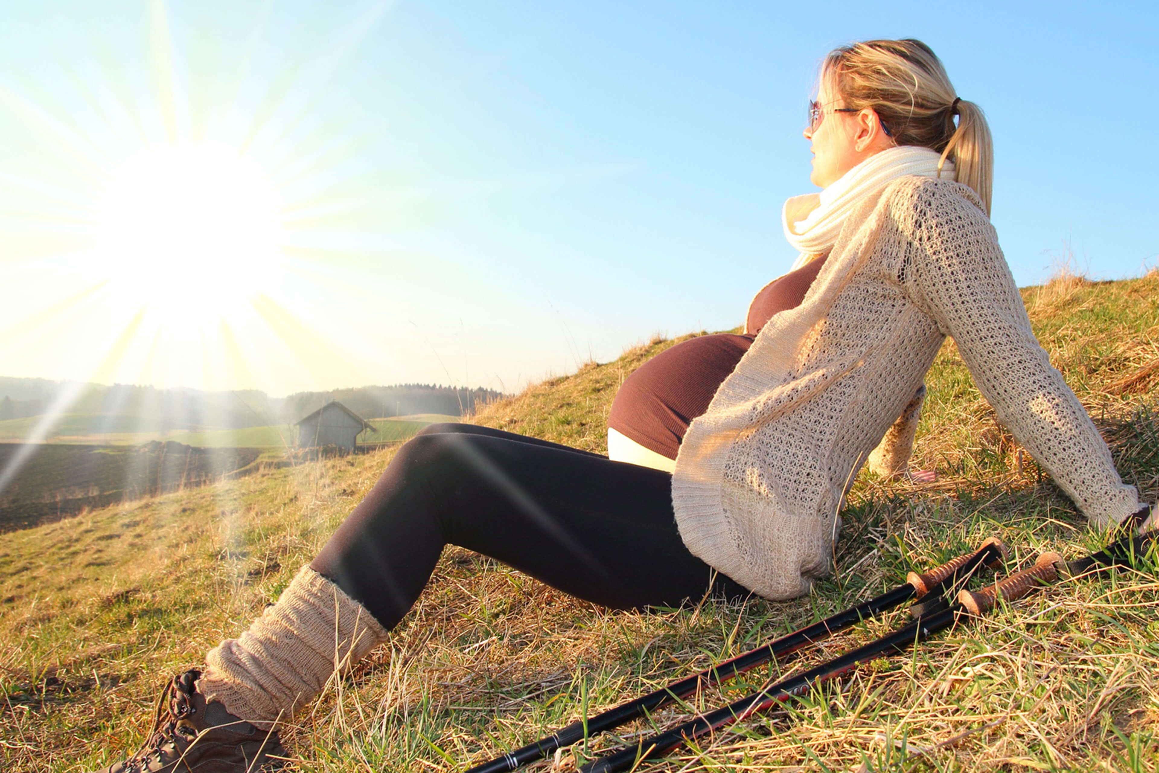 Eine Frau, die auf einem grasbewachsenen Hügel sitzt und in die Ferne blickt, vor einem sonnigen Hintergrund.