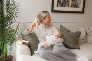 A woman lounges on a cream-colored couch, smiling while holding a sandwich in one hand and breastfeeding a baby nestled in her lap. A plate with a croissant and some raspberries rests on the arm of the sofa, and a leafy plant adds a cozy, homey touch to the scene