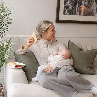 A woman lounges on a cream-colored couch, smiling while holding a sandwich in one hand and breastfeeding a baby nestled in her lap. A plate with a croissant and some raspberries rests on the arm of the sofa, and a leafy plant adds a cozy, homey touch to the scene.