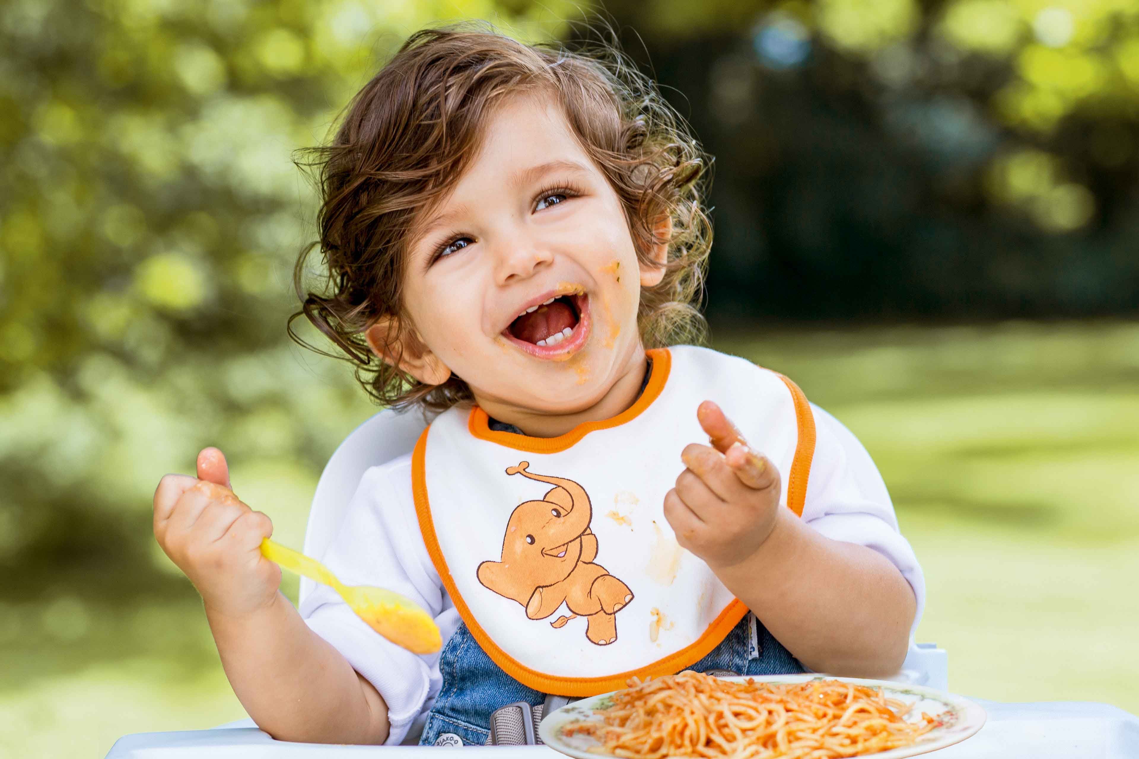 Ein fröhliches Kind, das im Freien sitzt und Spaghetti mit einem Lätzchen isst, auf dem ein Cartoon-Dinosaurier abgebildet ist.