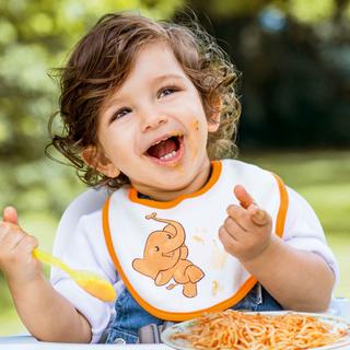 Ein fröhliches Kind, das im Freien sitzt und Spaghetti mit einem Lätzchen isst, auf dem ein Cartoon-Dinosaurier abgebildet ist.