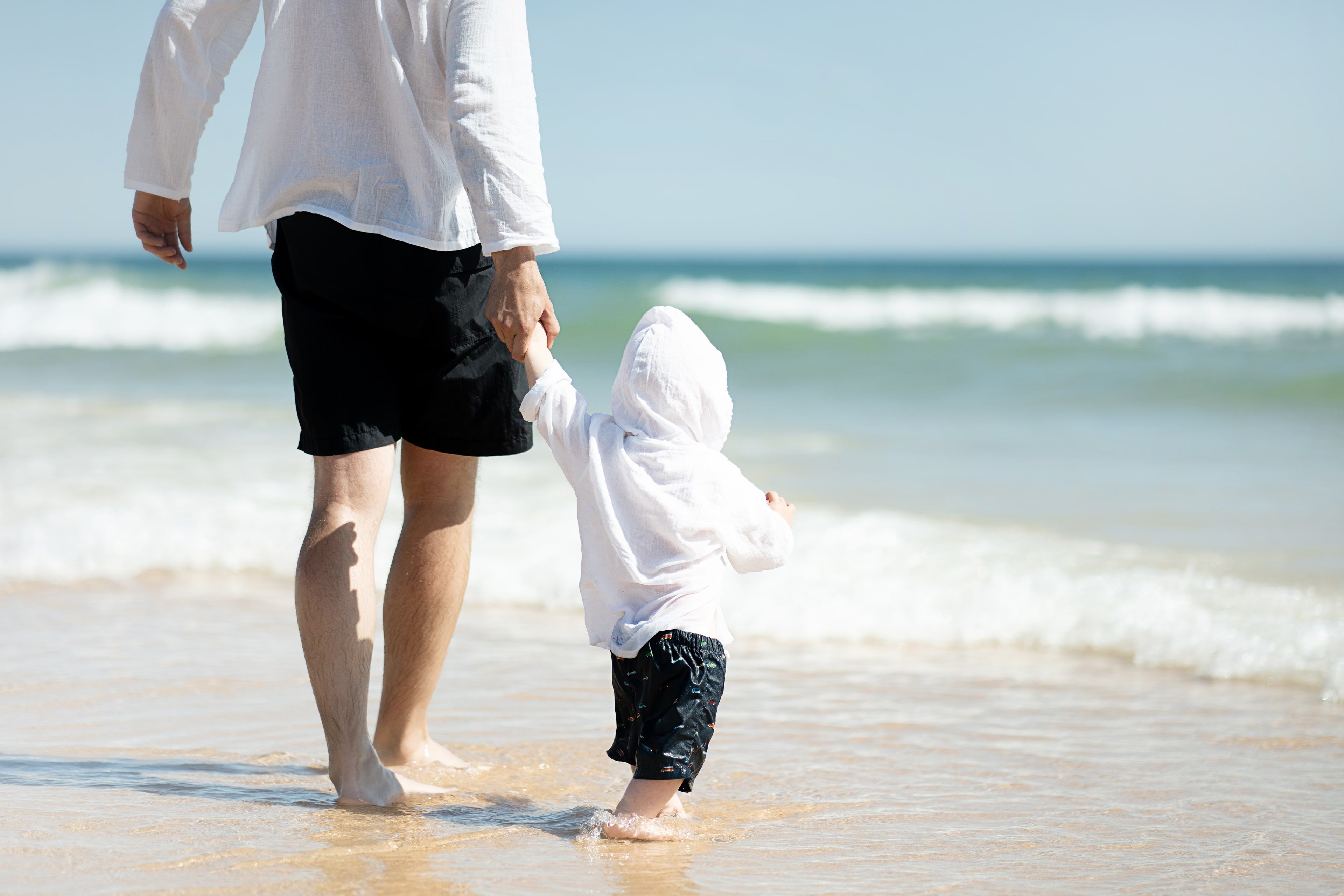 Eine heitere Strandszene, in der ein Erwachsener und ein Kind Hand in Hand am Ufer entlang spazieren, während im Hintergrund die Wellen sanft aneinanderschlagen.