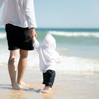 Eine heitere Strandszene, in der ein Erwachsener und ein Kind Hand in Hand am Ufer entlang spazieren, während im Hintergrund die Wellen sanft aneinanderschlagen.
