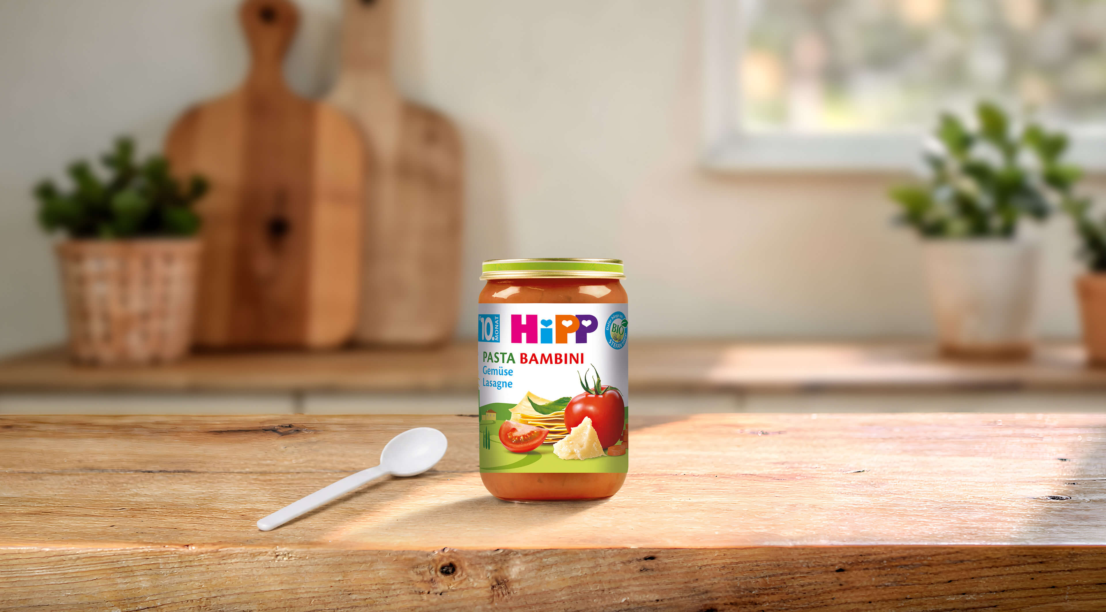 HiPP Pasta Bambini Gemüse-Lasagne Bio-Gläschen ab 10. Monat auf Holztisch mit Löffel in heller Küche
