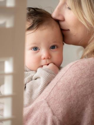 baby held in woman's arms, she seems to be smelling her baby's head and the baby is staring at the camera.