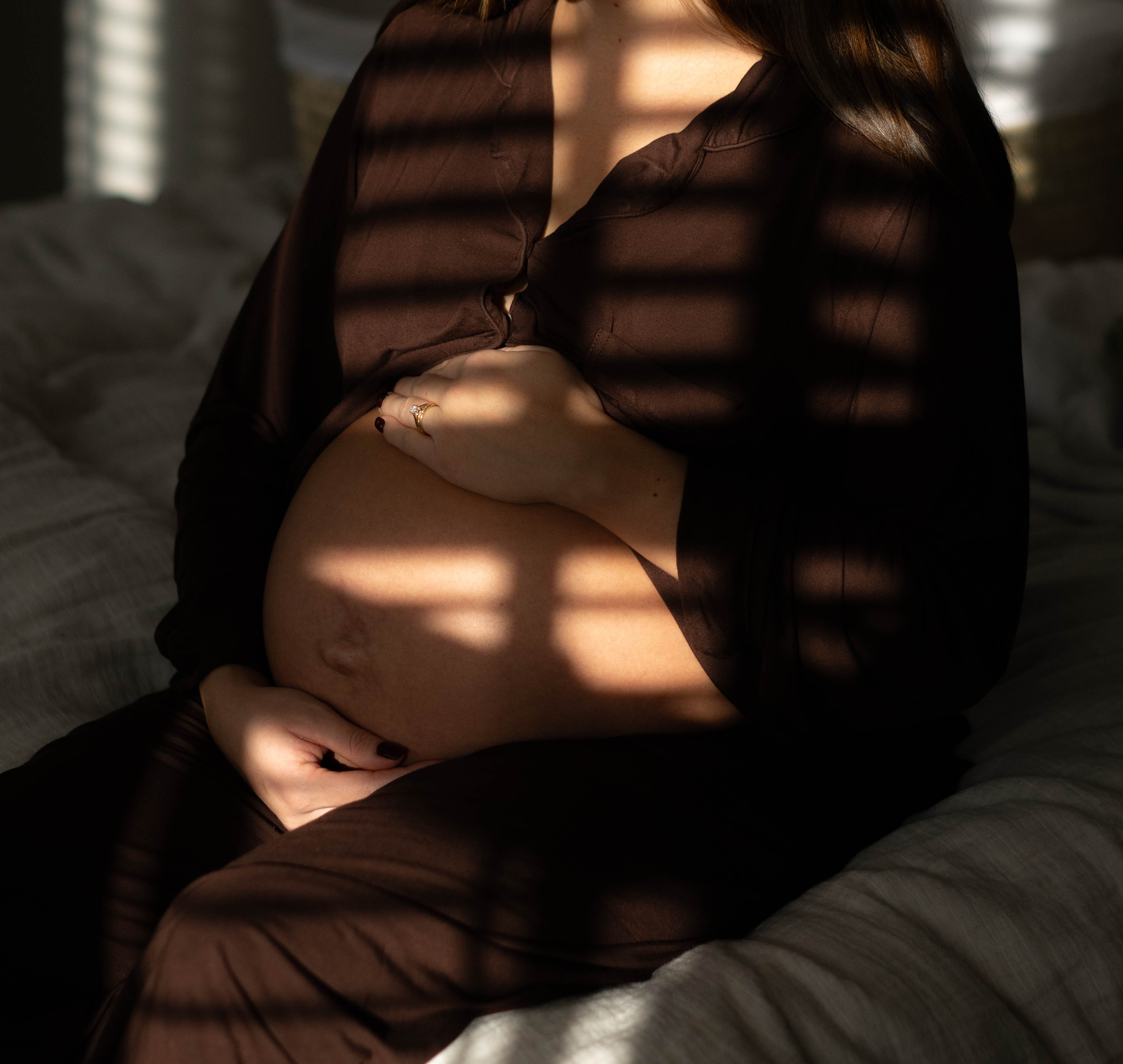 Heavily pregnant woman sitting on a bed, gently cradling her bump in soft, dappled light
