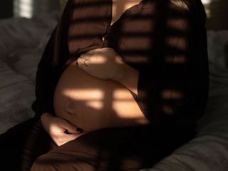 Heavily pregnant woman sitting on a bed, gently cradling her bump in soft, dappled light