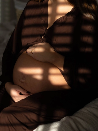 Heavily pregnant woman sitting on a bed, gently cradling her bump in soft, dappled light