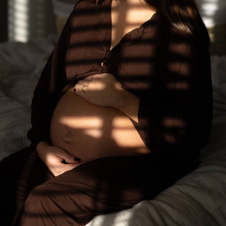 Heavily pregnant woman sitting on a bed, gently cradling her bump in soft, dappled light