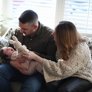 Dad holding upset baby whilst mum is trying to distract her with a teddy bear