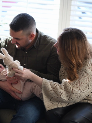 Dad holding upset baby whilst mum is trying to distract her with a teddy bear