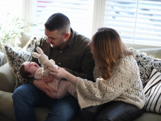 Dad holding upset baby whilst mum is trying to distract her with a teddy bear