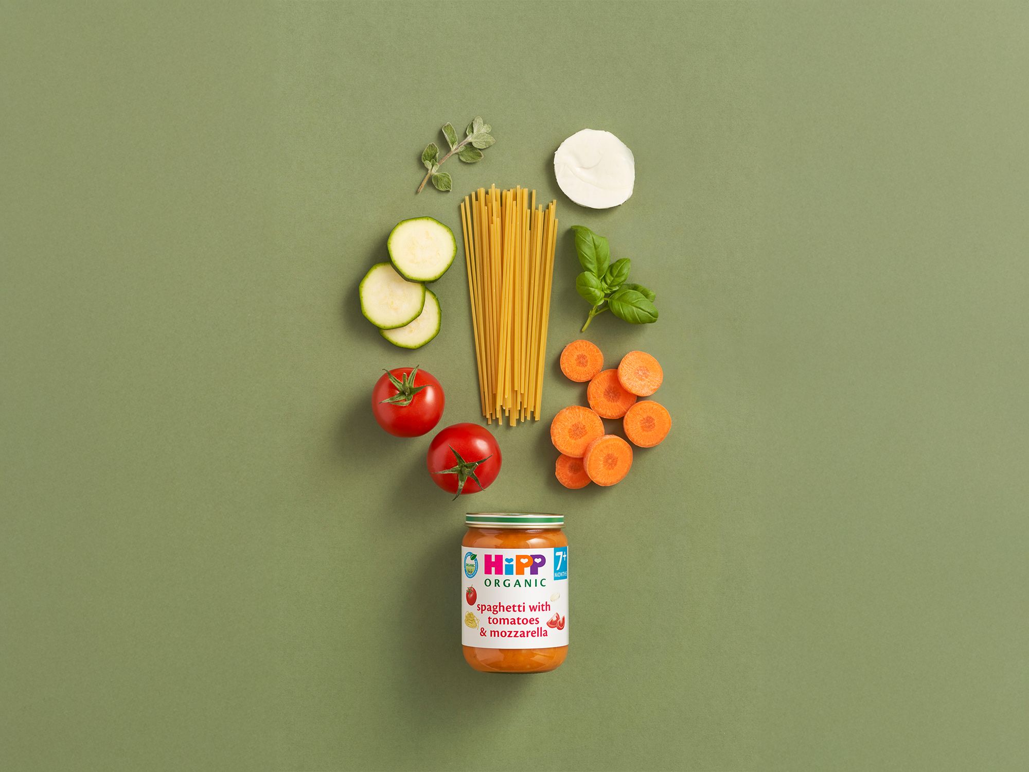 HiPP Organic Spaghetti with Tomatoes & Mozzarella jar surrounded by fresh tomatoes, courgette, carrots, spaghetti and herbs on green