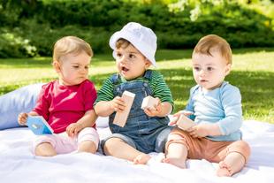 Drei Babys sitzen im Freien auf einer Decke, jedes hält ein Spielzeug, ein Baby trägt eine Mütze.