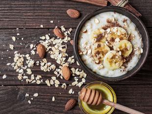 Eine Schüssel Haferflocken mit geschnittenen Bananen, Mandeln und Zimt, begleitet von einem Honiglöffel und gestreuten Haferflocken auf einem rustikalen Holzuntergrund.
