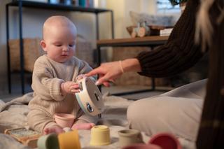 A baby in a cozy beige sweater sits on a soft blanket, holding a tambourine while an adult points at it, encouraging play. Colorful stacking cups and a xylophone are scattered nearby, creating a warm and playful learning moment