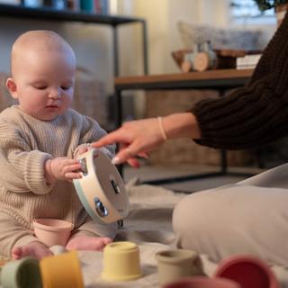 A baby in a cozy beige sweater sits on a soft blanket, holding a tambourine while an adult points at it, encouraging play. Colorful stacking cups and a xylophone are scattered nearby, creating a warm and playful learning moment.