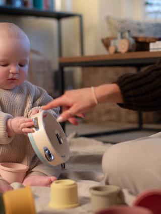 A baby in a cozy beige sweater sits on a soft blanket, holding a tambourine while an adult points at it, encouraging play. Colorful stacking cups and a xylophone are scattered nearby, creating a warm and playful learning moment.