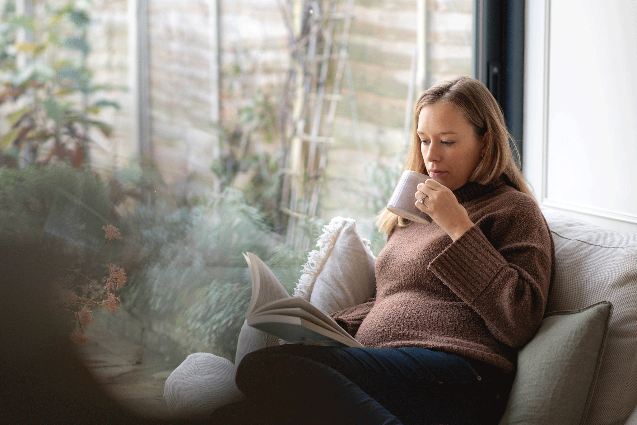 A pregnant woman sitting comfortably indoors on a sofa near a large garden window, reading a open book and holding a mug.