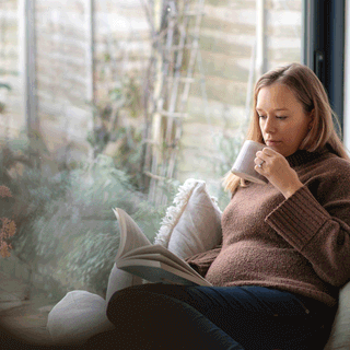 A pregnant woman sitting comfortably indoors on a sofa near a large garden window, reading a open book and holding a mug.