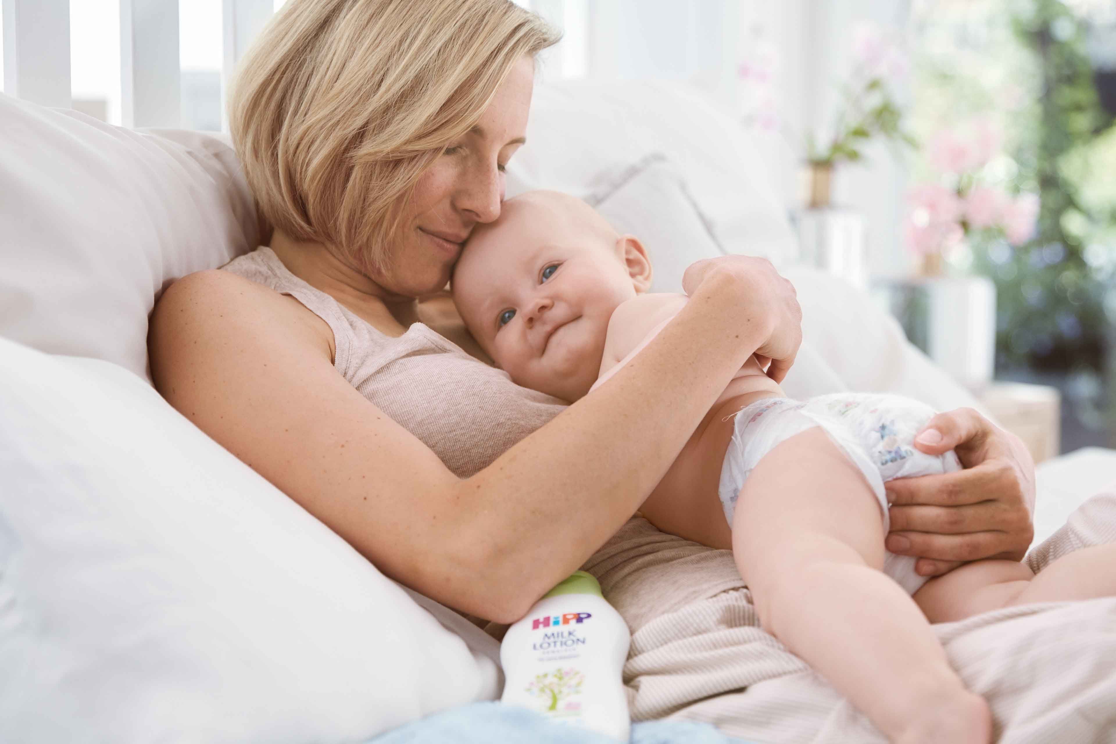 Mutter kuschelt entspannt mit ihrem lächelnden Baby im Bett, im Vordergrund steht HiPP Babysanft Pflegelotion für sensible Haut.