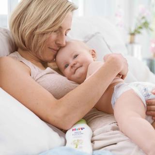Mutter kuschelt entspannt mit ihrem lächelnden Baby im Bett, im Vordergrund steht HiPP Babysanft Pflegelotion für sensible Haut.