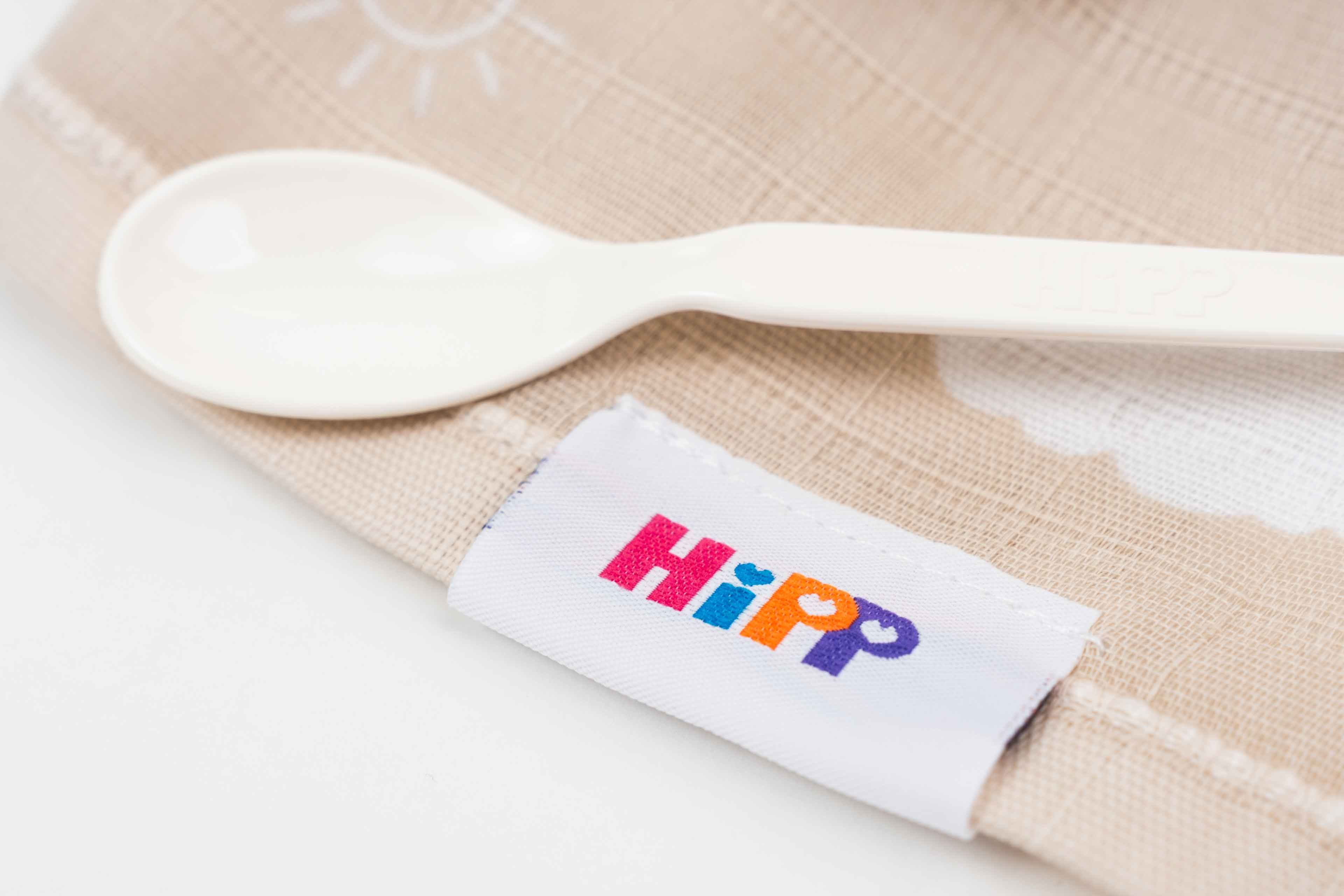 Ein weißer Babylöffel liegt auf einem hellen Stoff mit einem weißen Etikett, auf dem farbig 'HiPP' steht