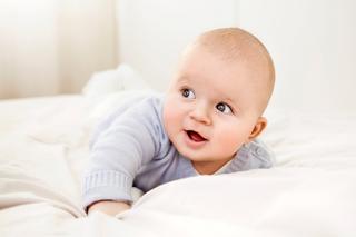 Lächelndes Baby in hellblauem Pulli liegt auf dem Bauch im Bett – Symbol für die Suche nach einem schönen Jungennamen