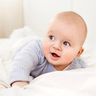 Lächelndes Baby in hellblauem Pulli liegt auf dem Bauch im Bett – Symbol für die Suche nach einem schönen Jungennamen