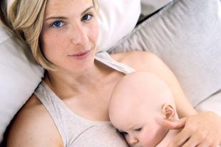 Eine Frau mit blondem Haar und blauen Augen, die ein Baby an ihrer Brust hält, beide liegen auf einer weichen Unterlage.
