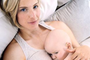 Eine Frau mit blondem Haar und blauen Augen, die ein Baby an ihrer Brust hält, beide liegen auf einer weichen Unterlage.