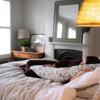 Pregnant woman resting on a cozy bed with a patterned body pillow, surrounded by soft bedding in a warmly lit bedroom.