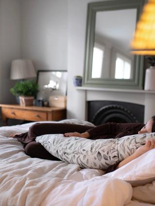 Pregnant woman resting on a cozy bed with a patterned body pillow, surrounded by soft bedding in a warmly lit bedroom.