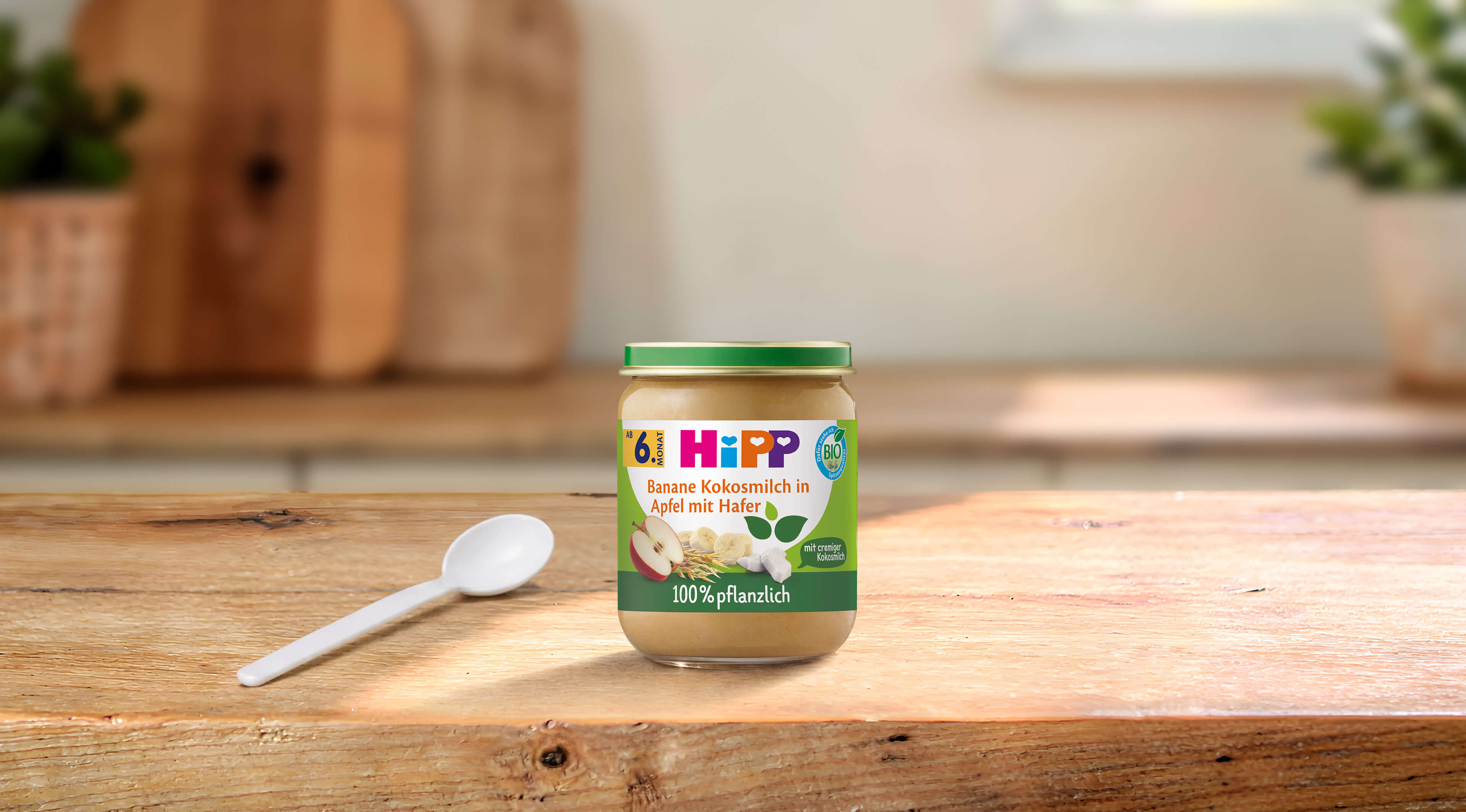 HiPP Bio-Gläschen Banane Kokosmilch in Apfel mit Hafer ab 6. Monat auf Holztisch mit Löffel in heller Küche