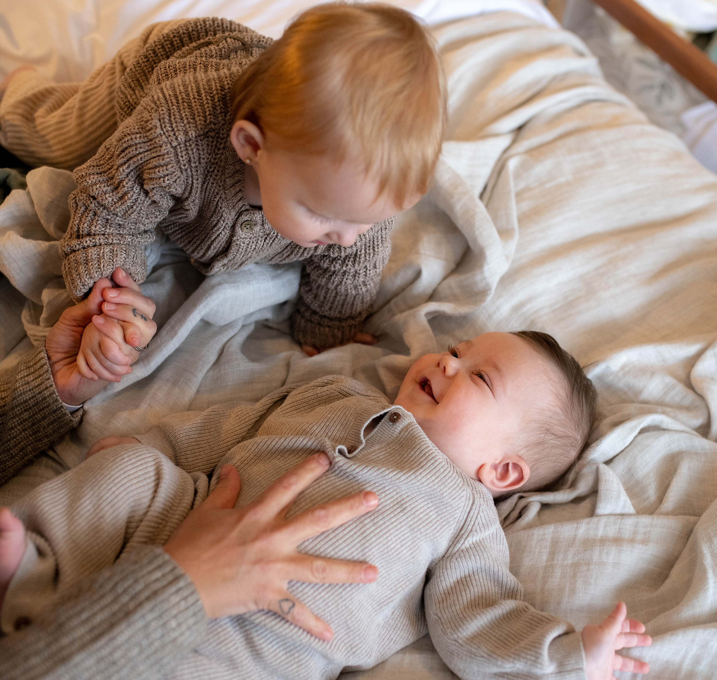 Two young children in cozy knitwear, one lying down and smiling, the other leaning over holding a hand, on a soft bed.