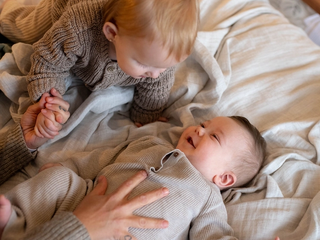 Two young children in cozy knitwear, one lying down and smiling, the other leaning over holding a hand, on a soft bed.