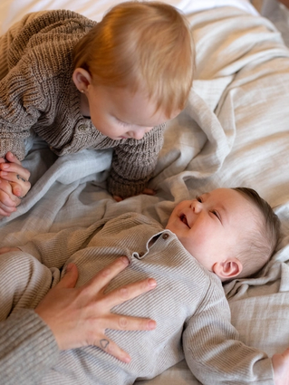 Two young children in cozy knitwear, one lying down and smiling, the other leaning over holding a hand, on a soft bed.