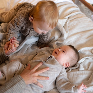 Two young children in cozy knitwear, one lying down and smiling, the other leaning over holding a hand, on a soft bed.