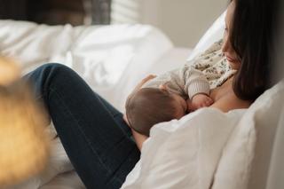 Mother breastfeeding her baby in bed, holding them close while resting against soft white pillows.
