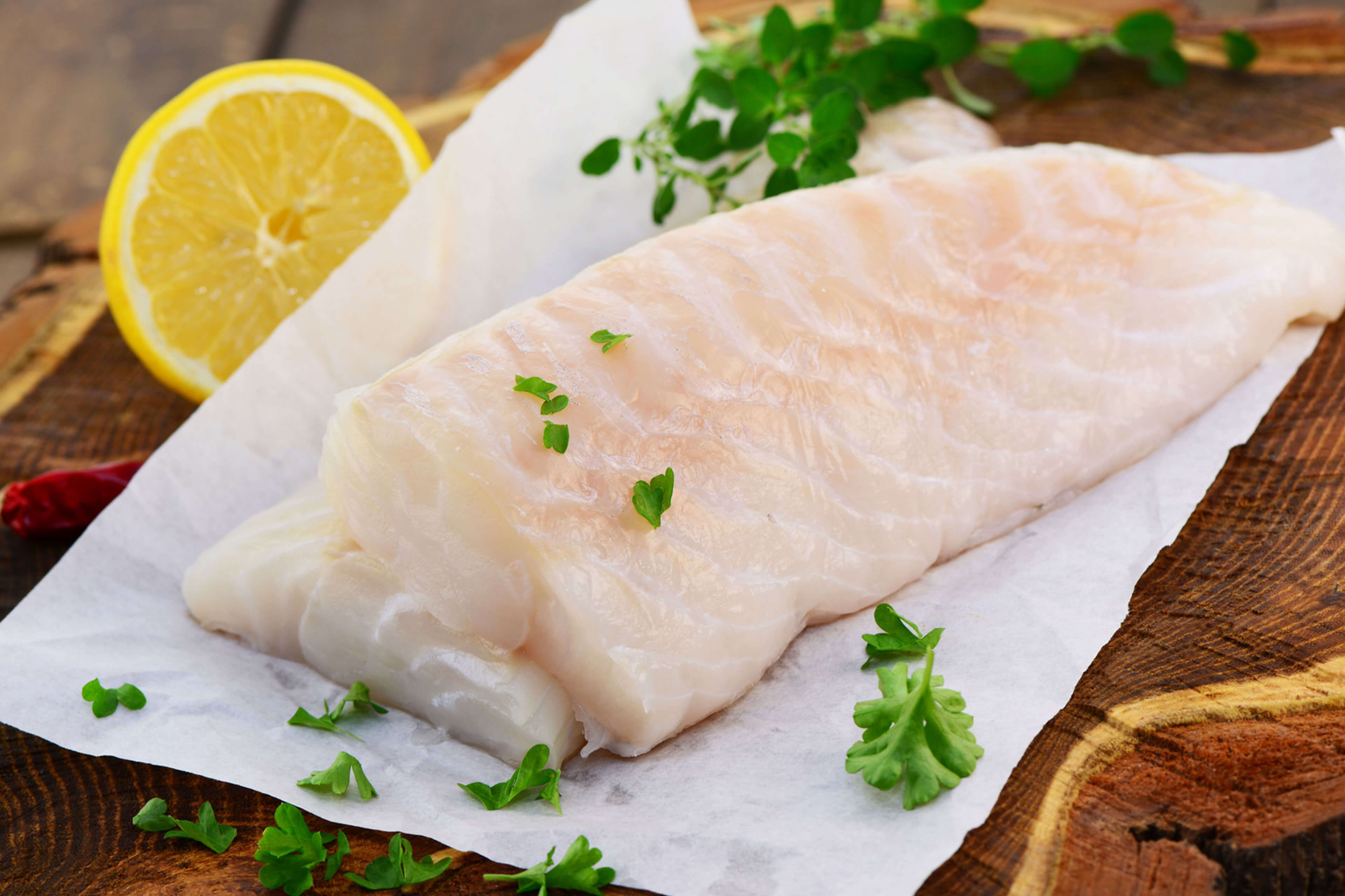 Ein Stück roher Fisch auf weißem Papier, garniert mit grünen Kräutern und einer Zitronenscheibe, auf einer Holzunterlage platziert.