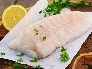 Ein Stück roher Fisch auf weißem Papier, garniert mit grünen Kräutern und einer Zitronenscheibe, auf einer Holzunterlage platziert.