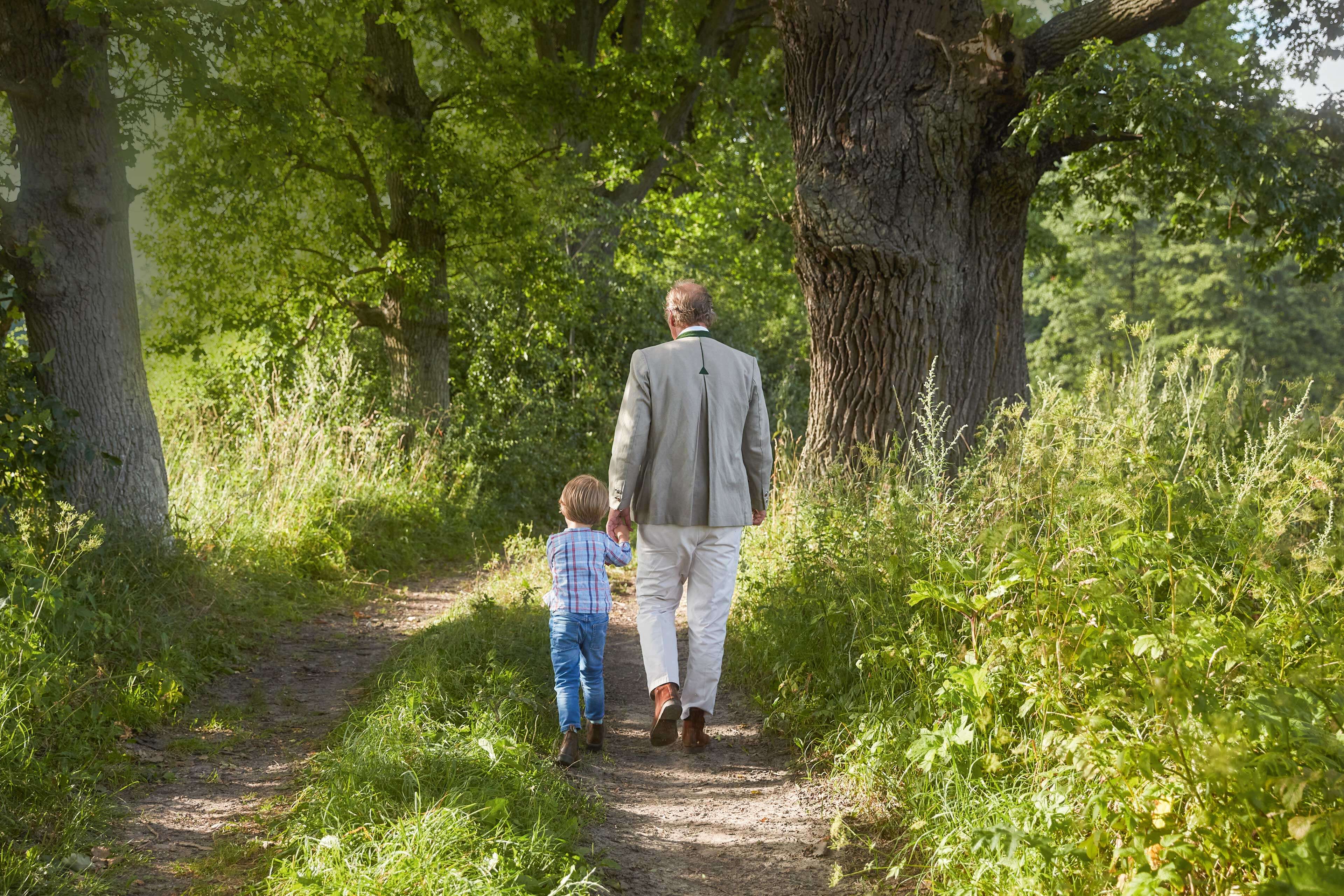Erwachsener und Kind gehen Hand in Hand auf einem naturbelassenen Waldweg, umgeben von alten Bäumen und viel Grün.