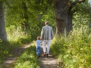 Erwachsener und Kind gehen Hand in Hand auf einem naturbelassenen Waldweg, umgeben von alten Bäumen und viel Grün.