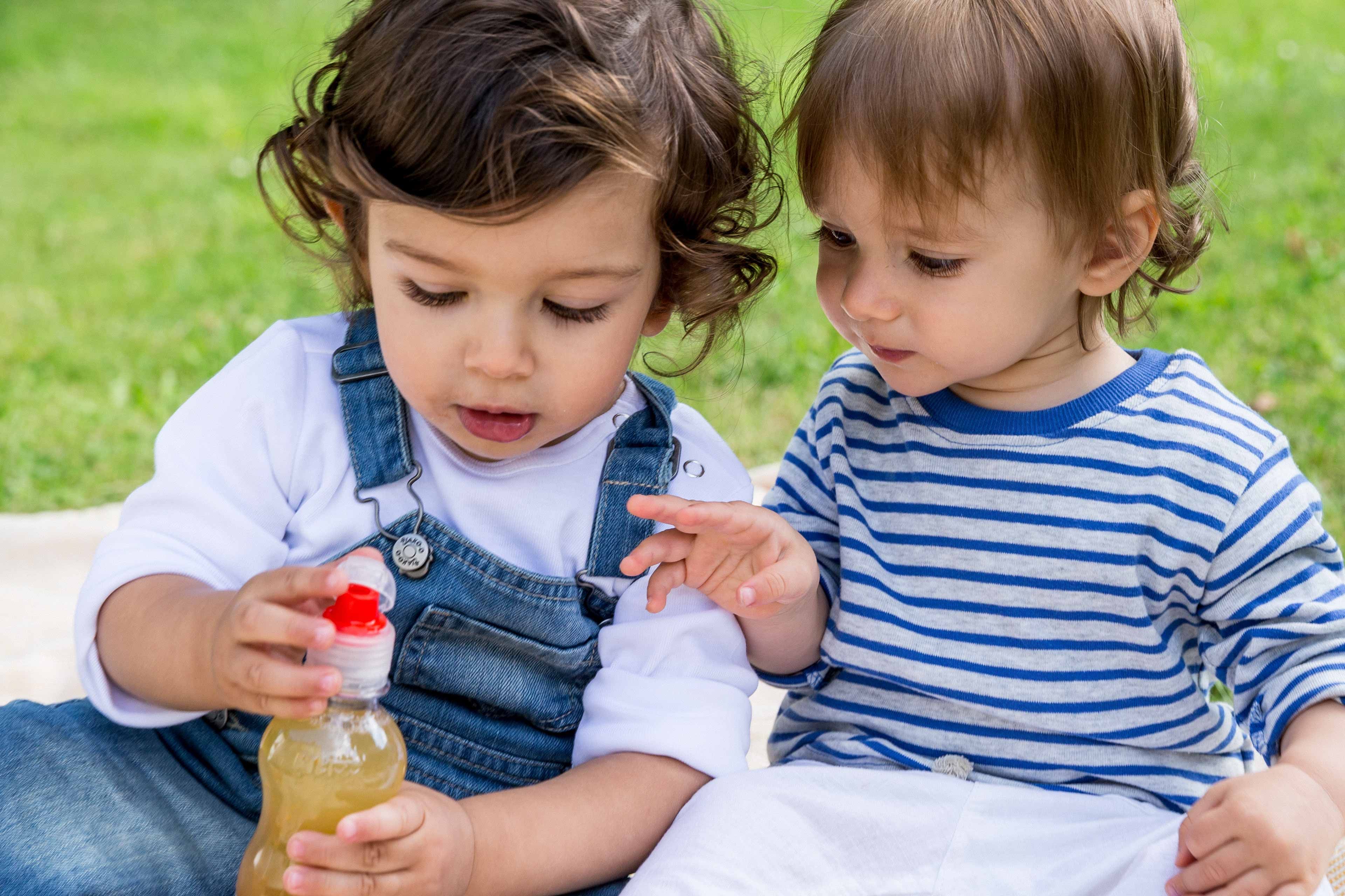 Zwei kleine Kinder, eines in weißem Hemd und blauer Latzhose, das andere in einem gestreiften Hemd, sitzen auf einer Decke auf einer Wiese und betrachten aufmerksam eine Flasche Saft.