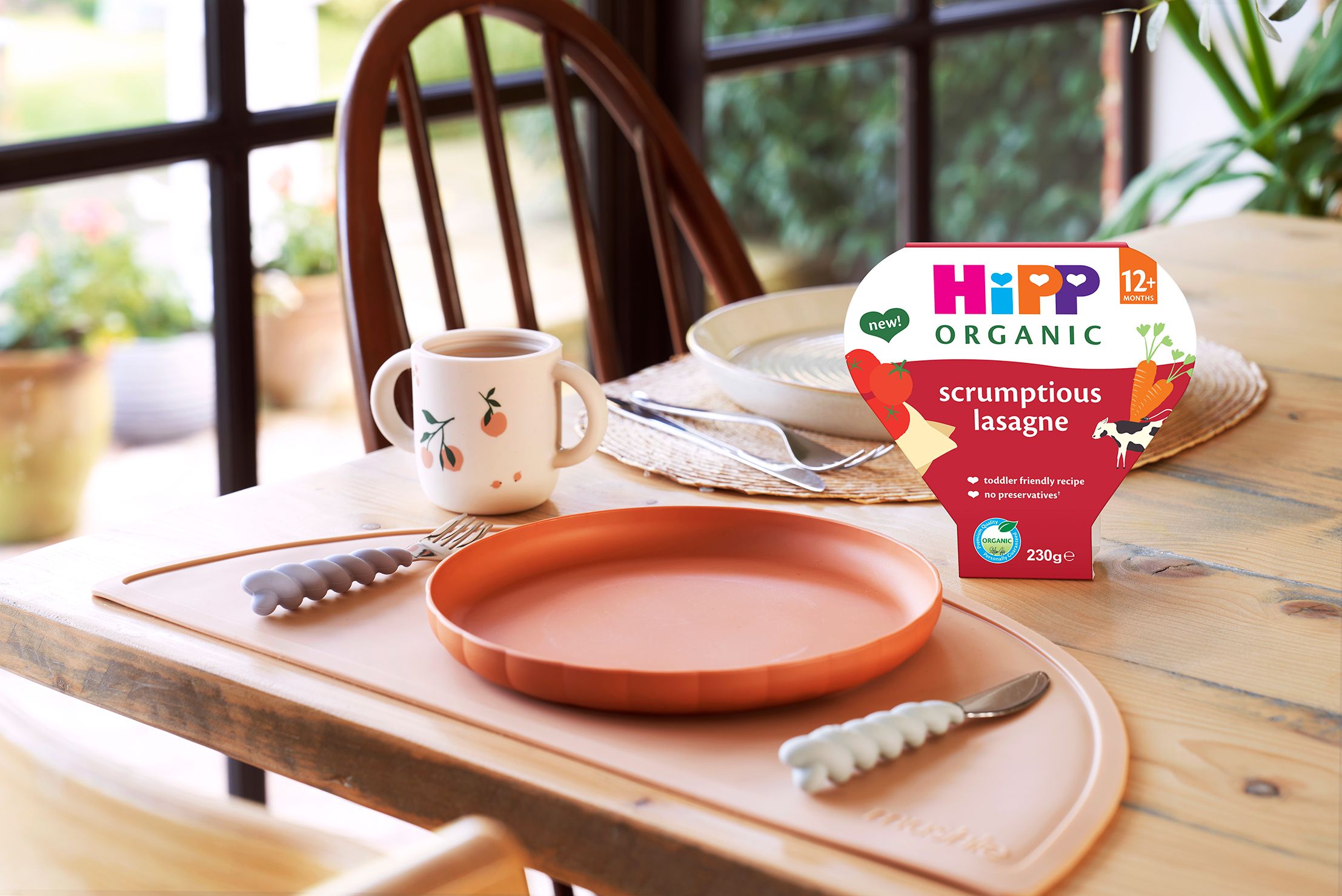 HiPP Organic Scrumptious Lasagne tray meal on a wooden dinning table, next to a toddlers plate, cutlery and cup