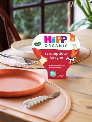 HiPP Organic Scrumptious Lasagne tray meal on a wooden dinning table, next to a toddlers plate, cutlery and cup